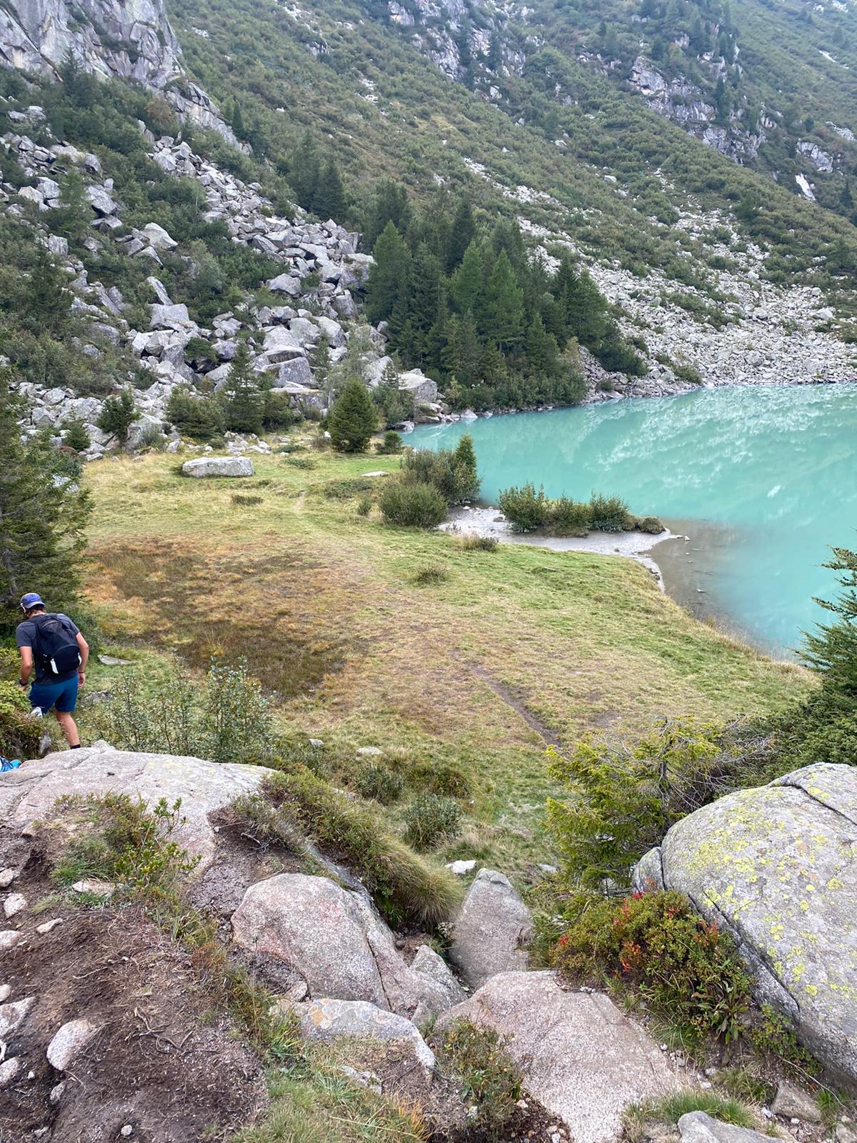 Vista di montagna ricoperta di verde con alcuni alberi dietro ad un lago cristallino ed un ragazzo che sta scendendo da delle grandi rocce