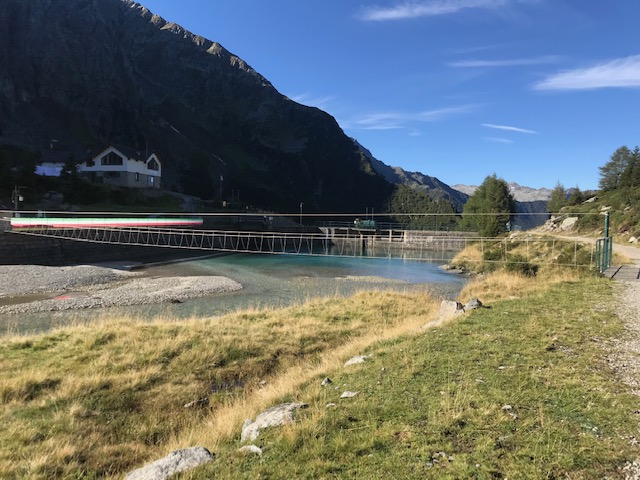 Foto di un ponte sopra un lago contornato da montagne e dagli alberi che riporta ad un rifugio