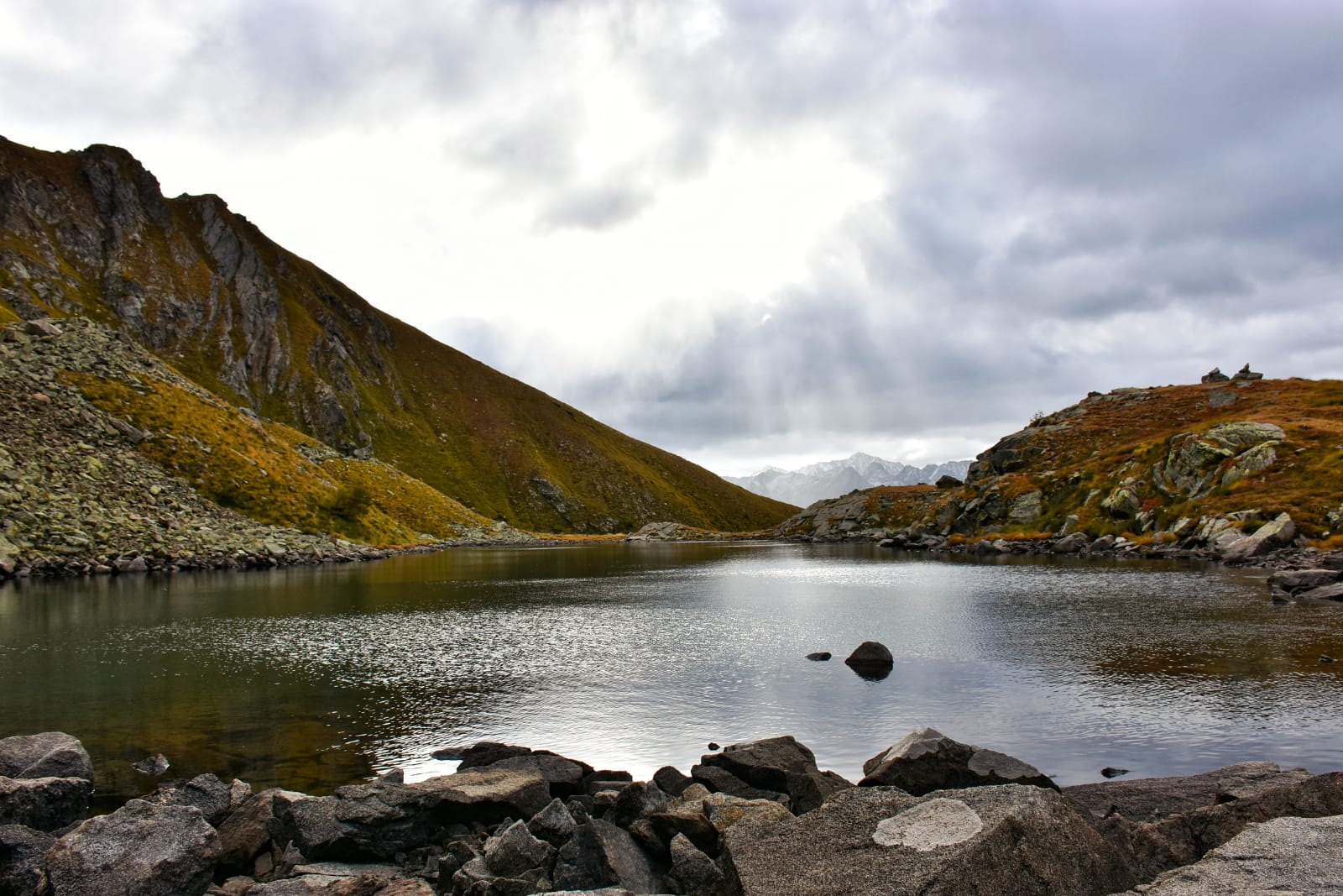 Vista lago contornato da grandi rocce e basse montagne rivestite di piante appassite su uno sfondo di un cielo grigio e cupo