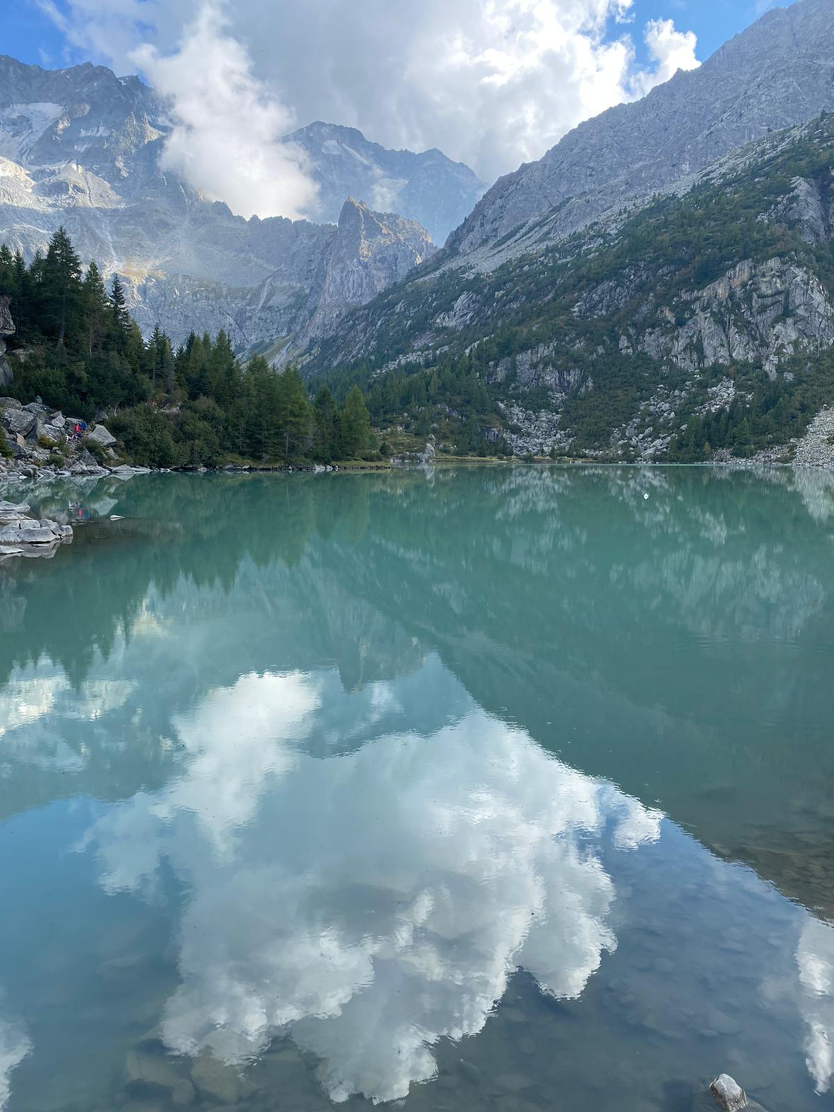 Vista di un lago cristallino circondato da rocce e alberi di fronte a delle montagne alti che si perdono tra le nuvole di un cielo sereno