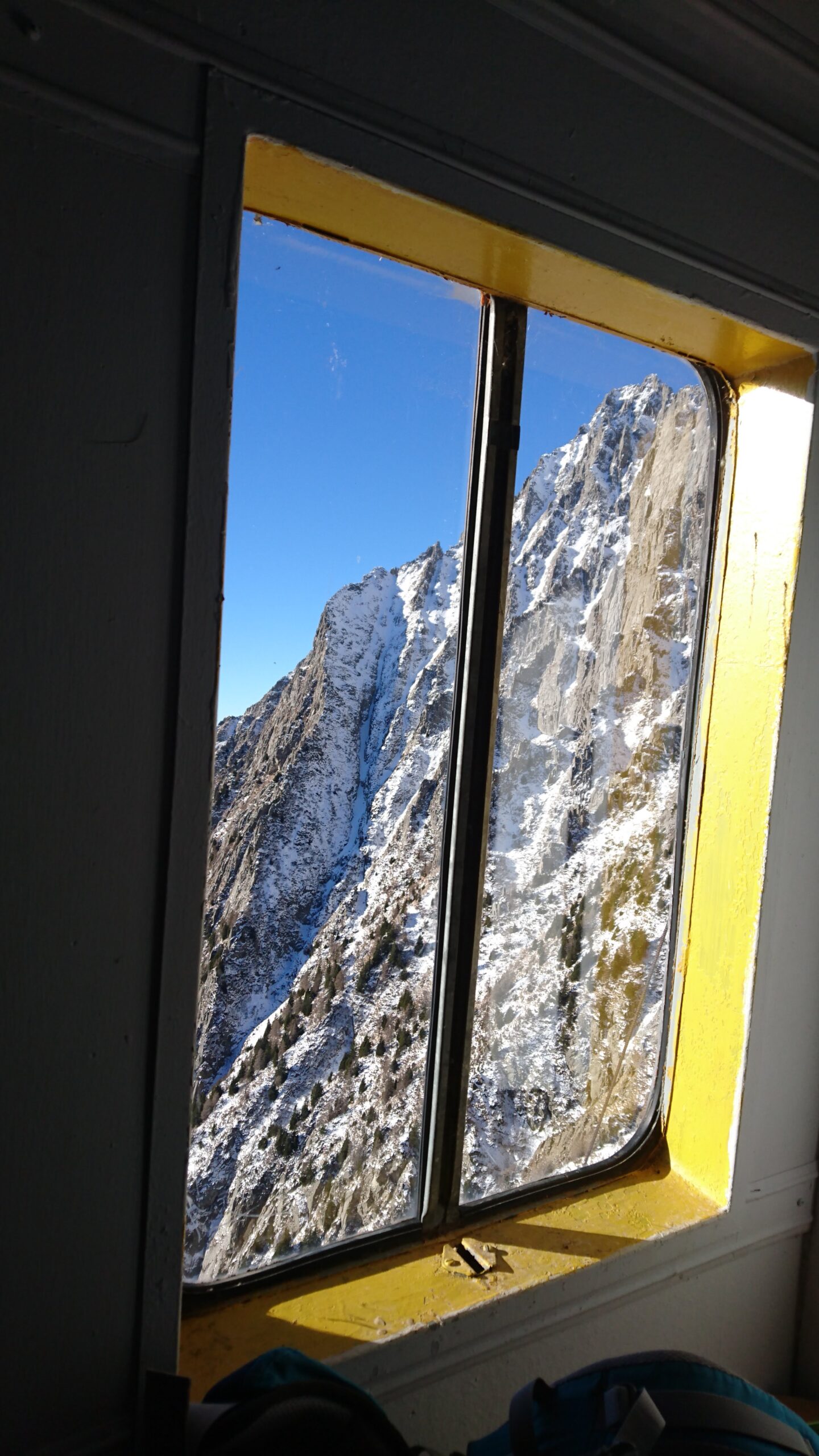 Foto di una finestra di un rifugio che si affaccia su una montagna con chiazze di neve