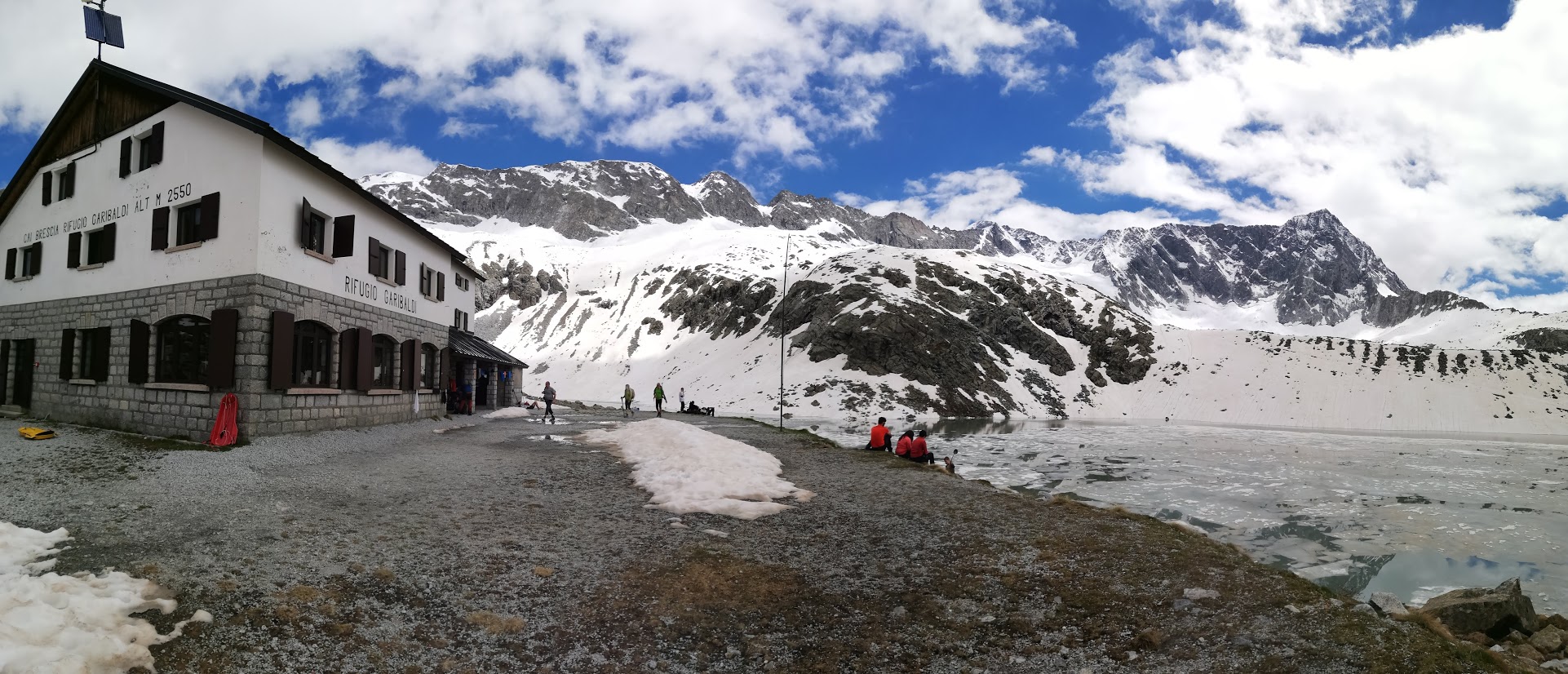 Foto in primo piano di un rifugio alpino con delle persone al di fuori e in secondo piano delle montagne innevate