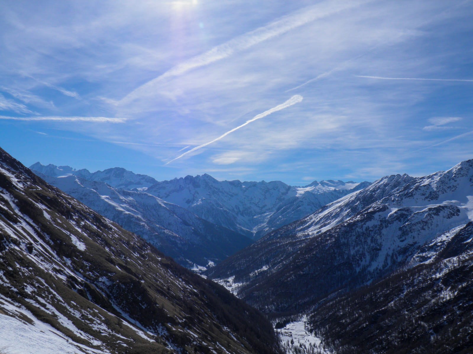 Vista di un cielo sereno e soleggiato con piccole nuvole sparse, in primo piano ci sono delle montagne scure e ricoperte di neve