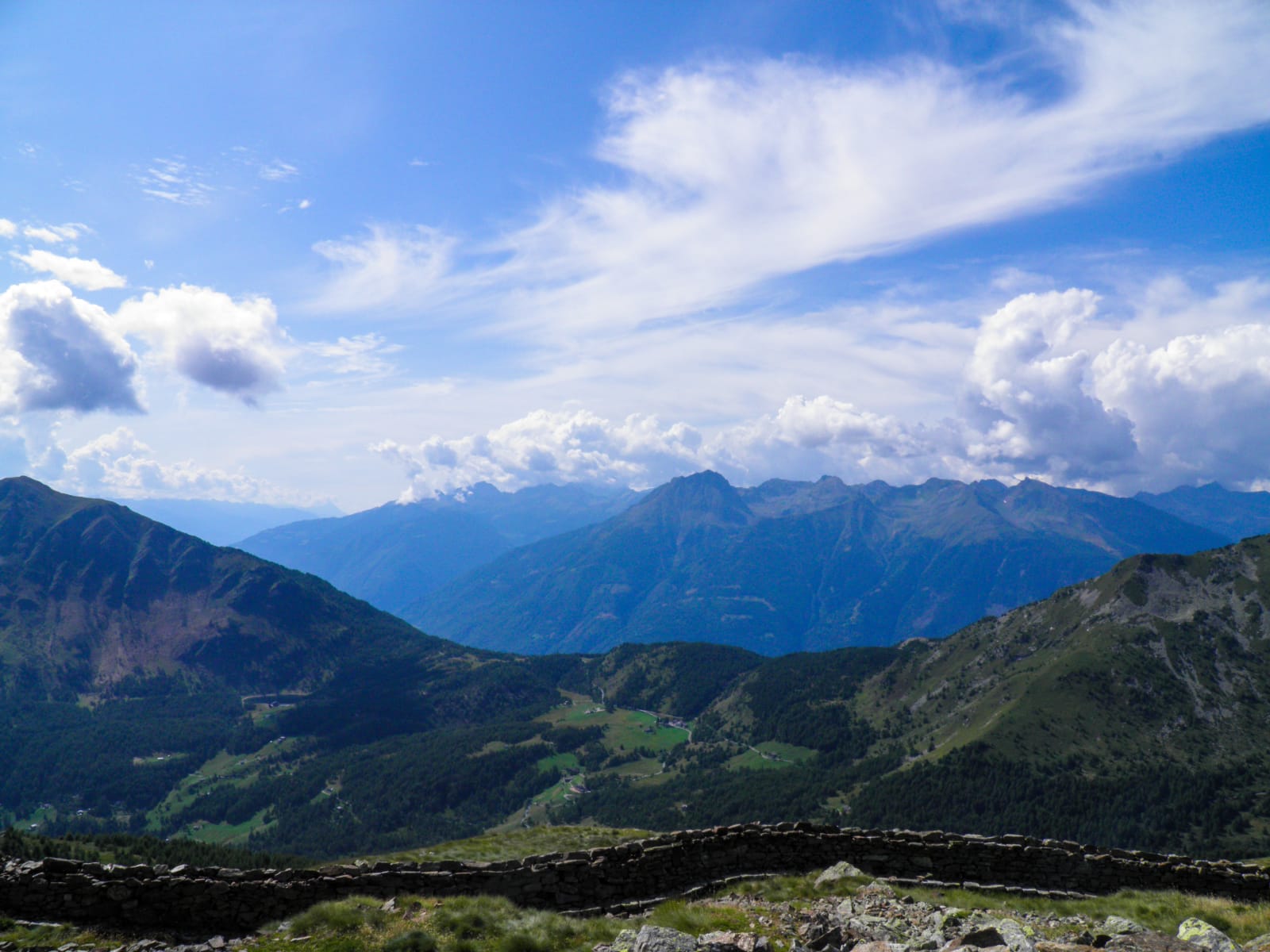 Vista di un piccolo sentiero recinto da basse muraglie in roccia di fronte ad una distesa di montagne ricoperte dal verde e da alberi