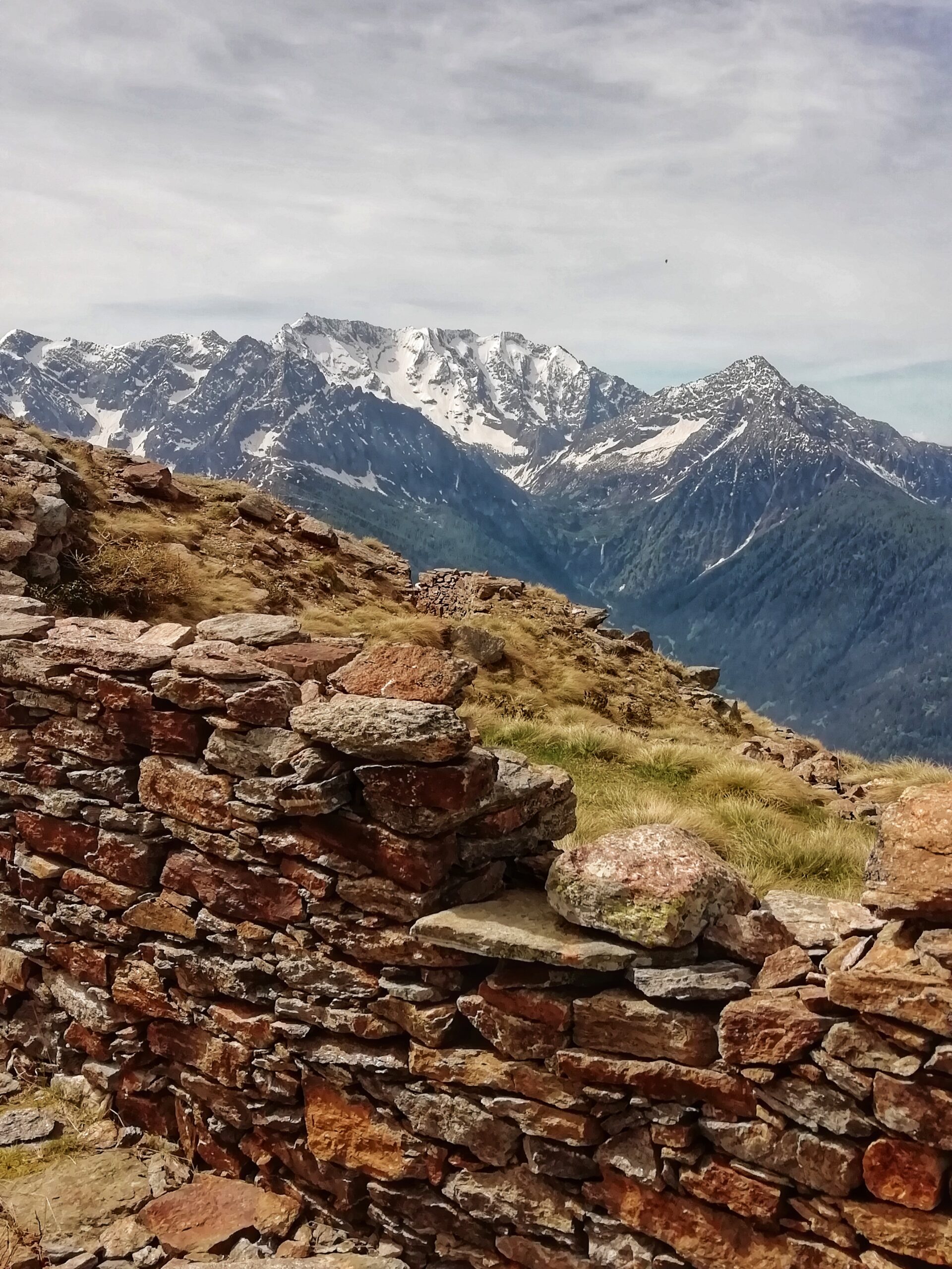 Vista di una piccola muraglia fatta di rocce con in secondo piano la vista delle montagne con piccole chiazze di neve e un cielo grigiastro