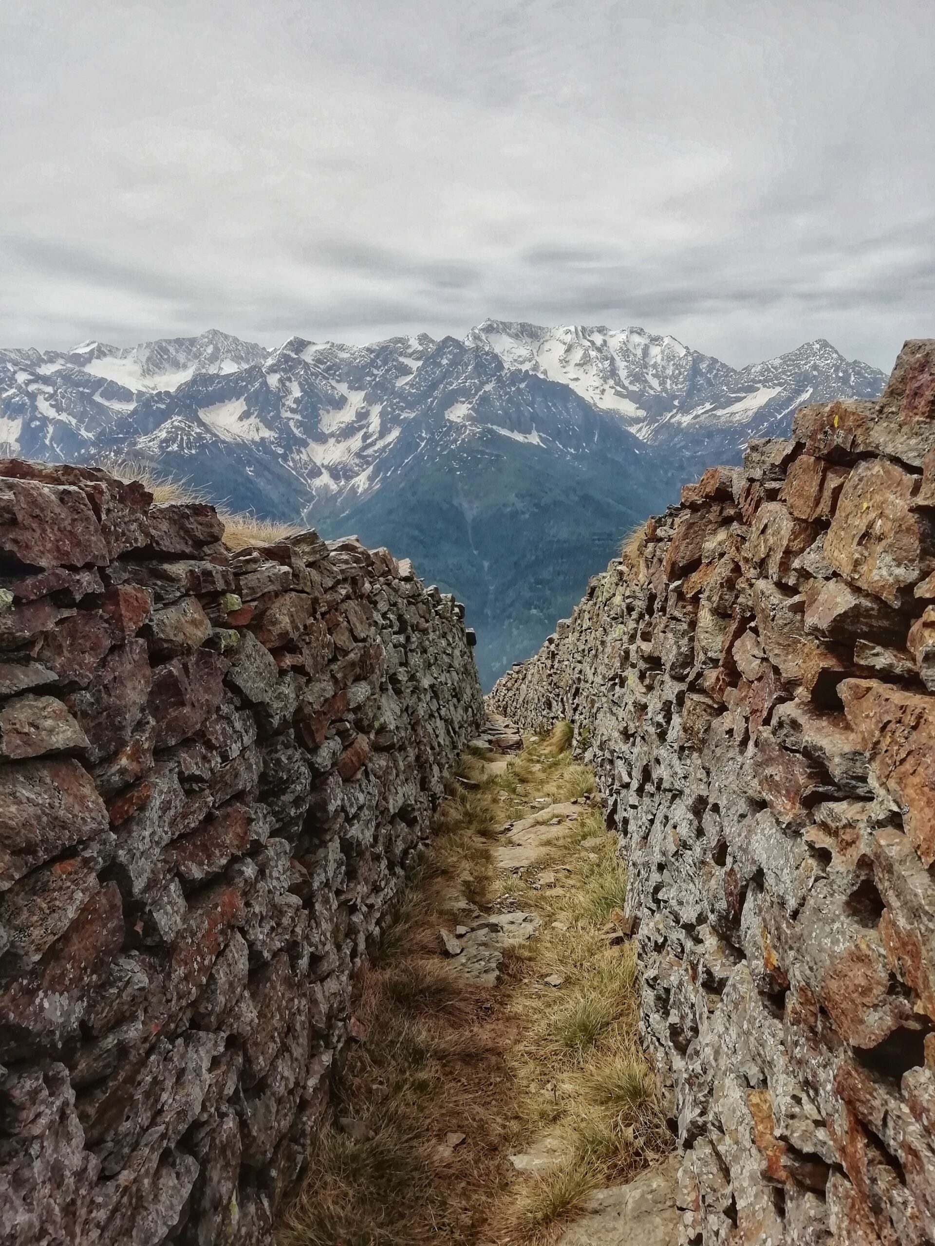 Vista montagne con chiazze di neve, da un sentiero con pareti in roccia e un cielo offuscato