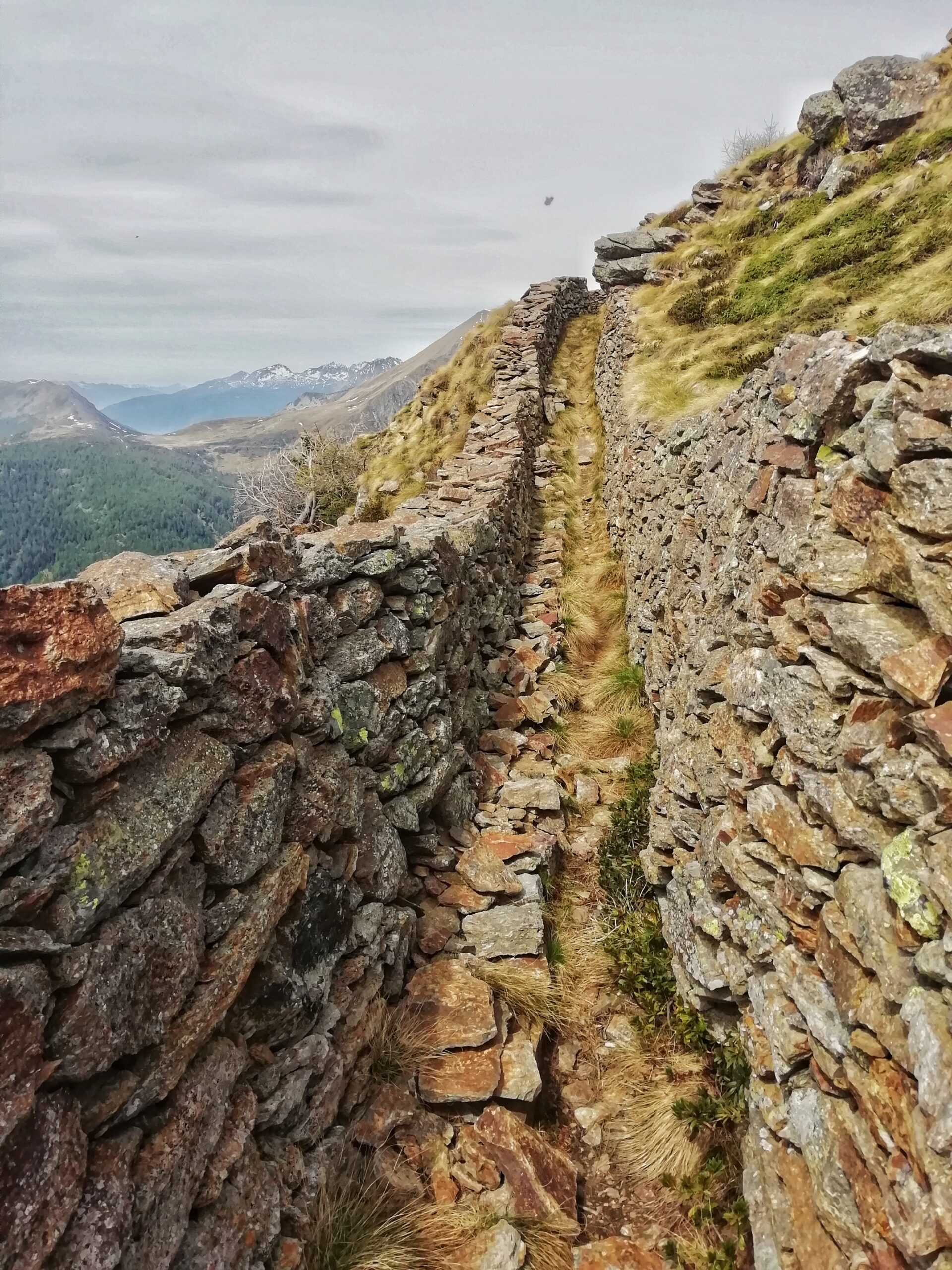 Foto rappresenta un sentiero stretto e costeggiato da una muraglia fatta di rocce in montagna