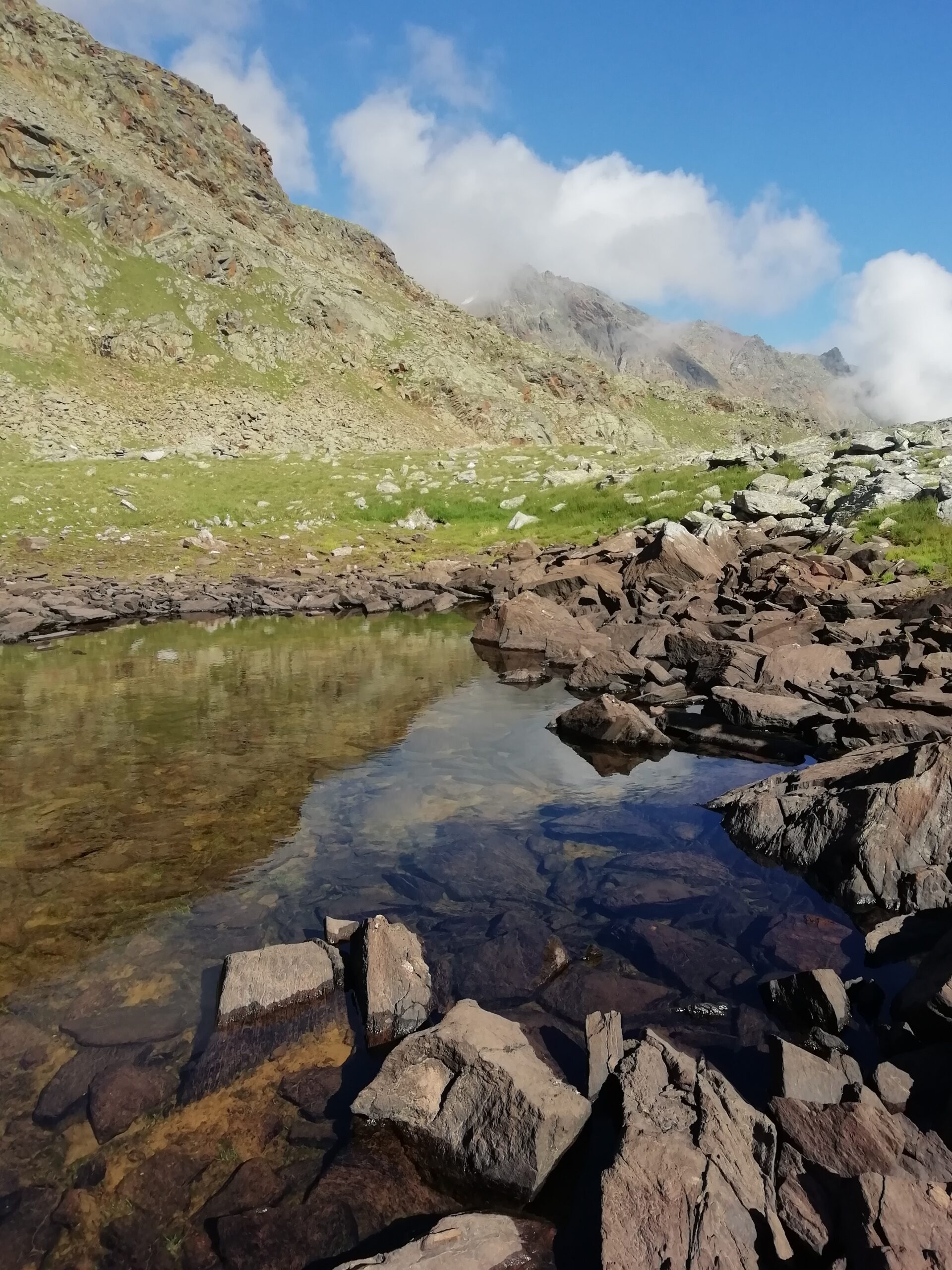 Vista lago con grandi rocce, a ridosso di montagne e un cielo nitido e poche nuvole