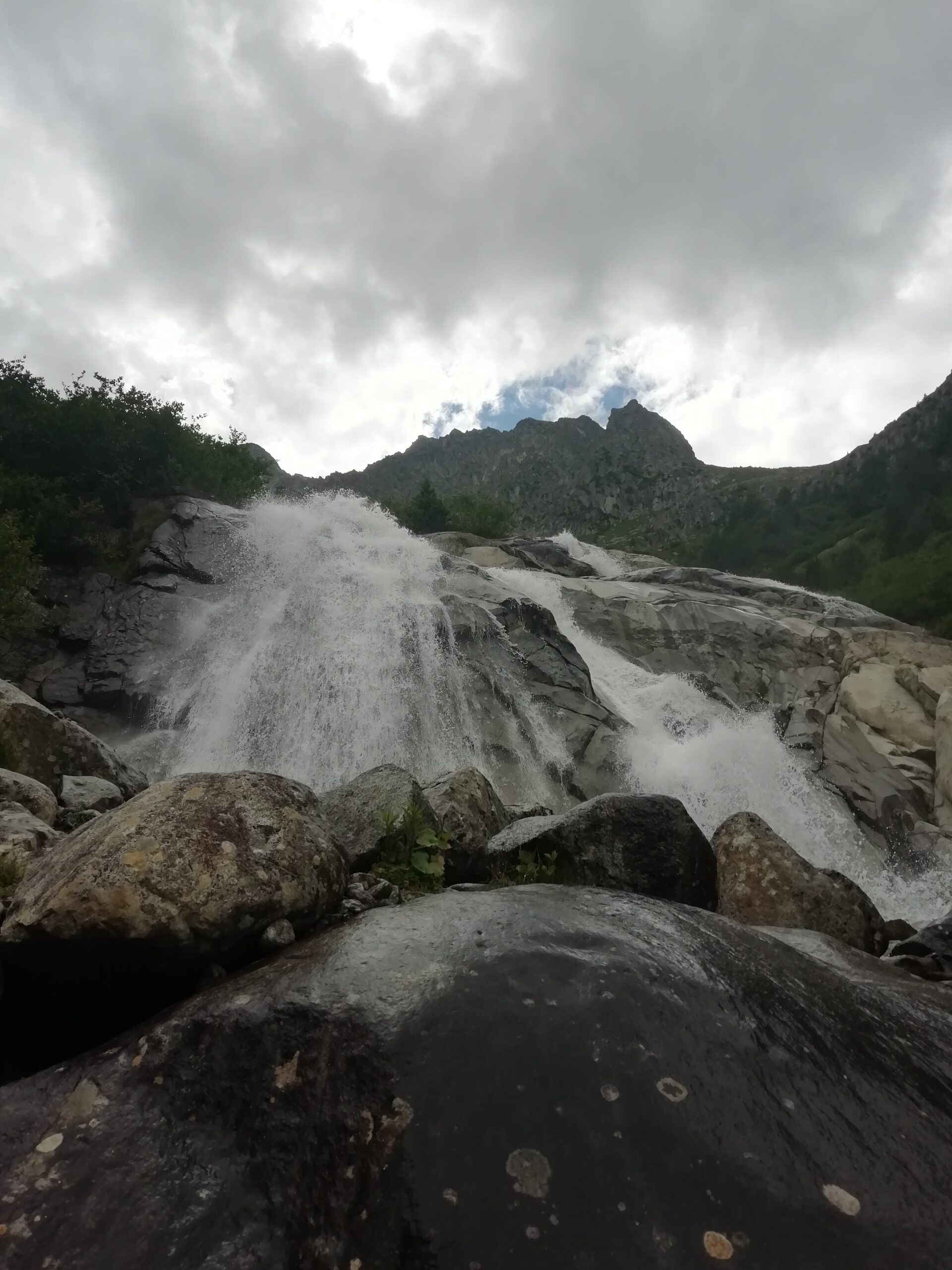 Foto di una cascata con acqua gelida, in sottofondo delle montagne scure e un cielo nuvoloso