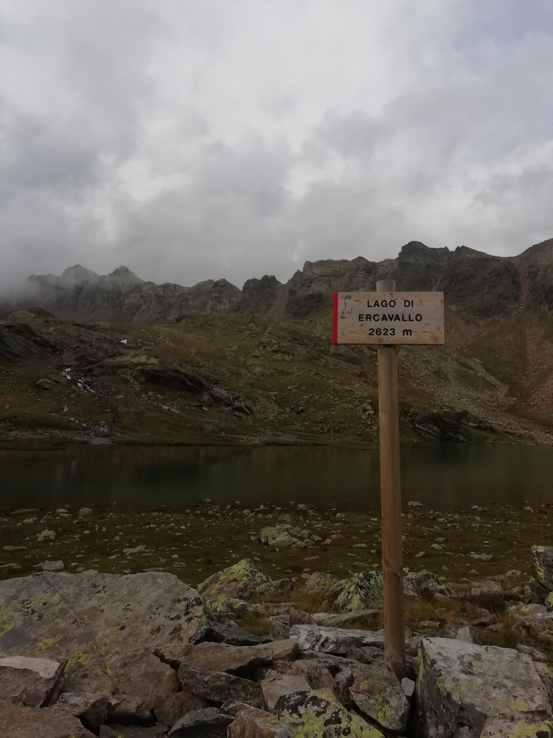 La foto riprende un cartello del Lago di Ercavallo posizionato in mezzo a delle rocce su uno sfondo del lago stesso e delle sue montagne che lo accompagnano, il tutto contornato con un cielo nuvoloso